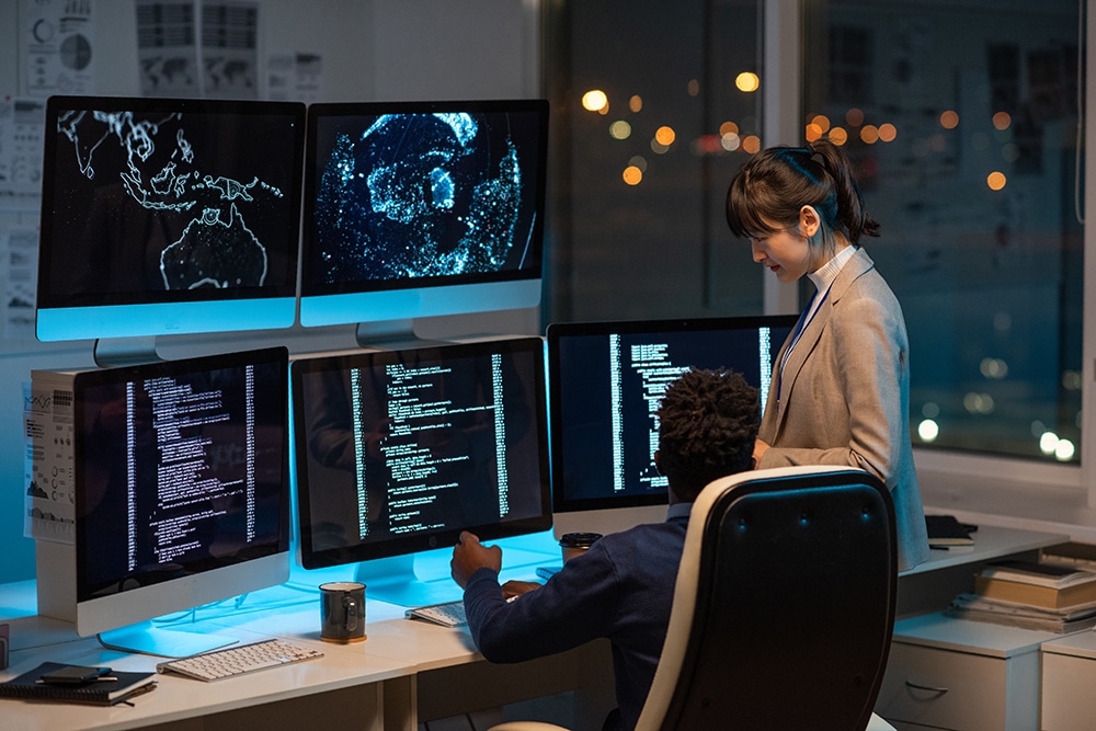 Two intercultural programmers consulting about working points while decoding data on computer screen in office